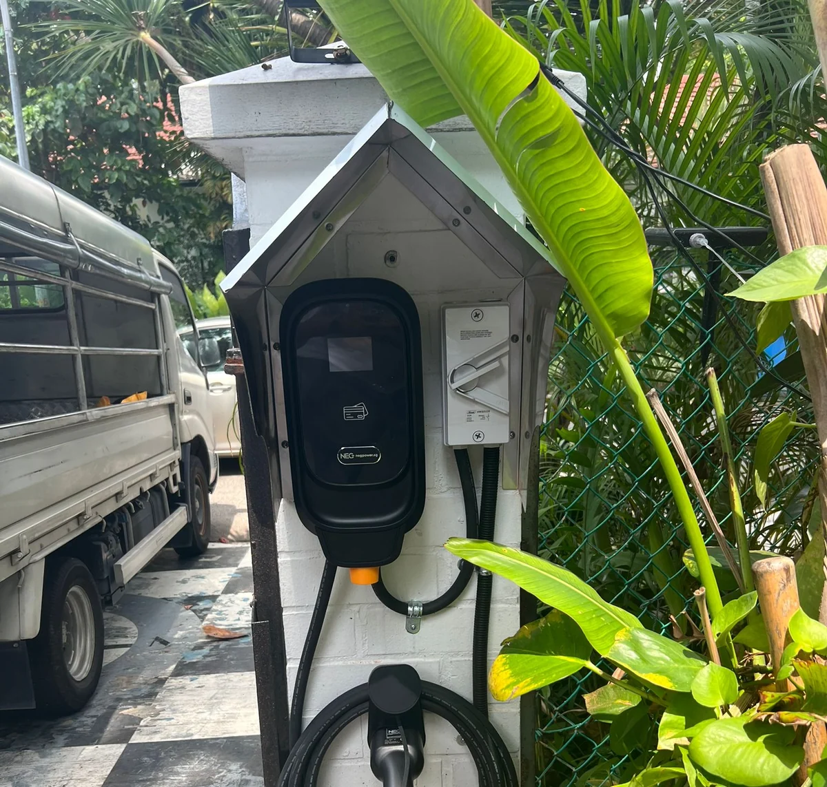 A black NEG charger under a small pitched weather hood on a wall, an isolator beside it, with banana leaves overhanging and a lorry parked alongside.