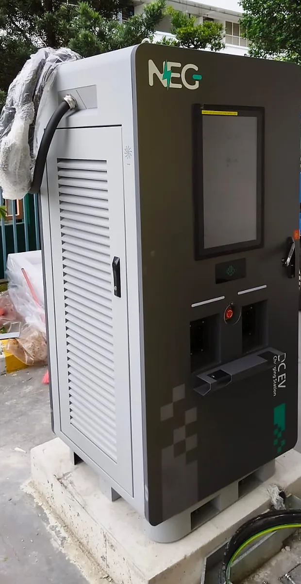 A NEG DC fast charger seen whole from the front corner — louvred cooling flank, colour screen, twin outlets and emergency stop — standing on a concrete plinth.