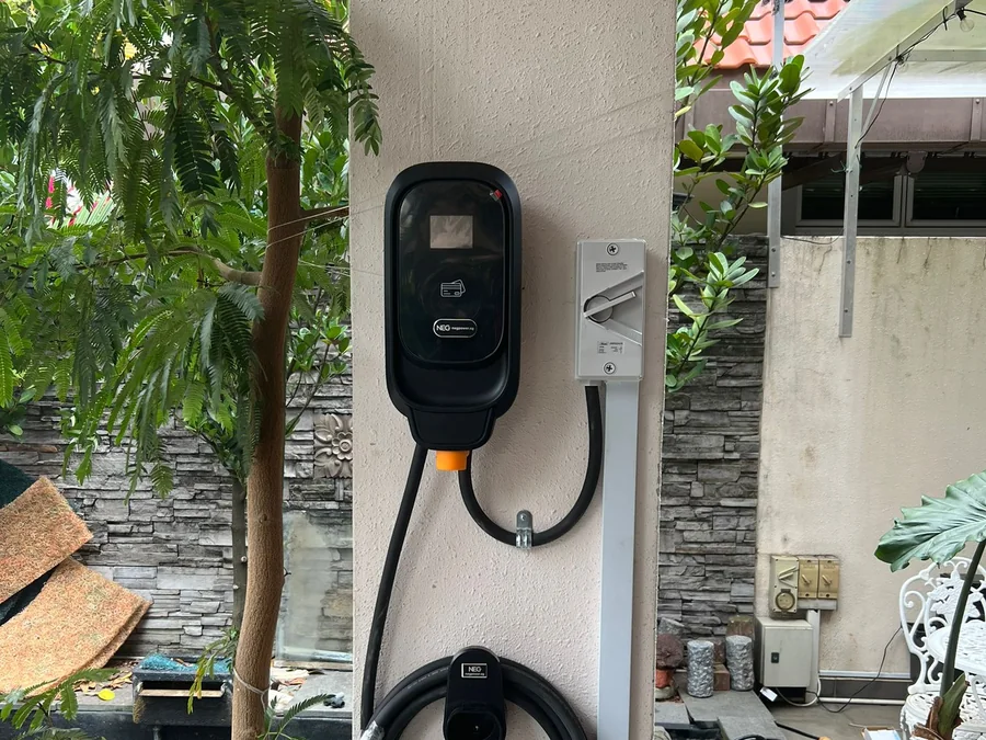 A black NEG wall charger on an exterior wall beside planting, the supply entering through conduit and the cable coiled on its holster.