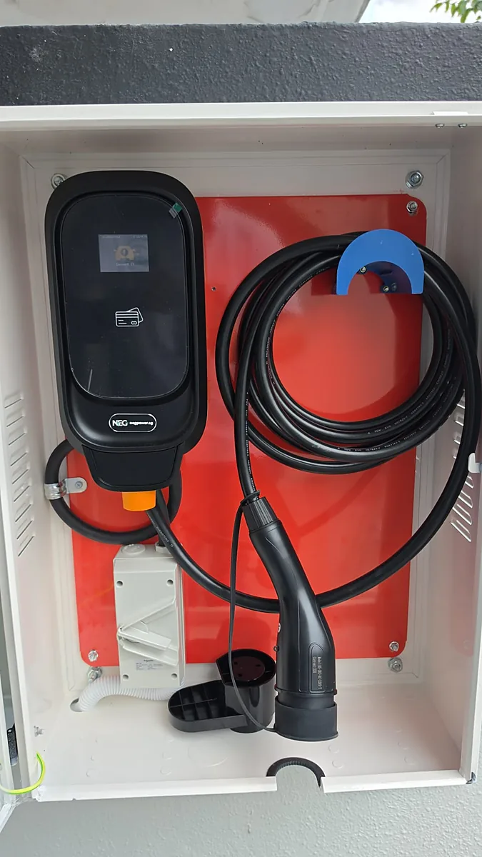 A black EV charger with a lit display, mounted on a red backboard inside a white louvred outdoor cabinet, its coiled lead hung on a blue hook with the connector hanging beside it.