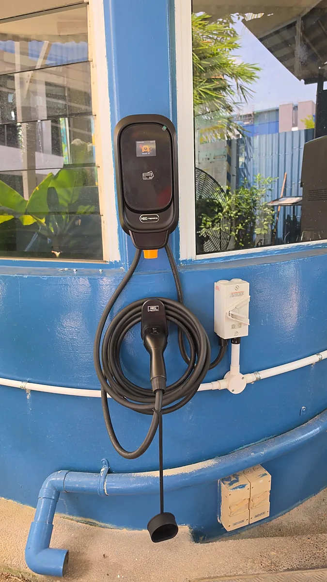 A black wall-mounted EV charger on a curved blue wall, its coiled lead and connector hanging below and a weatherproof isolator with white conduit alongside.