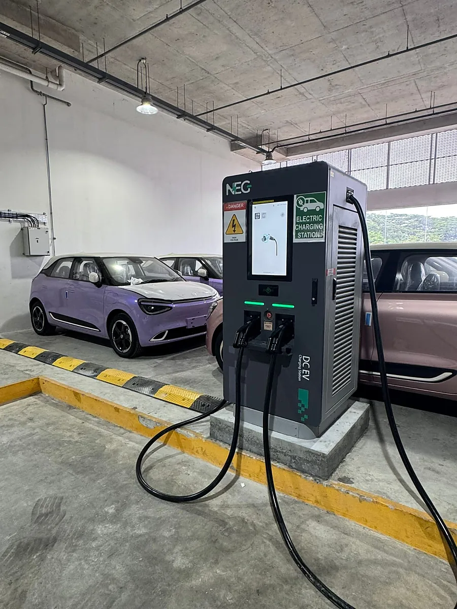 A free-standing DC charging cabinet on a concrete plinth in a covered car park, a red high-voltage label and a green "Electric Charging Station" sign beside its touchscreen, its charging cables running loose across a yellow-lined bay.