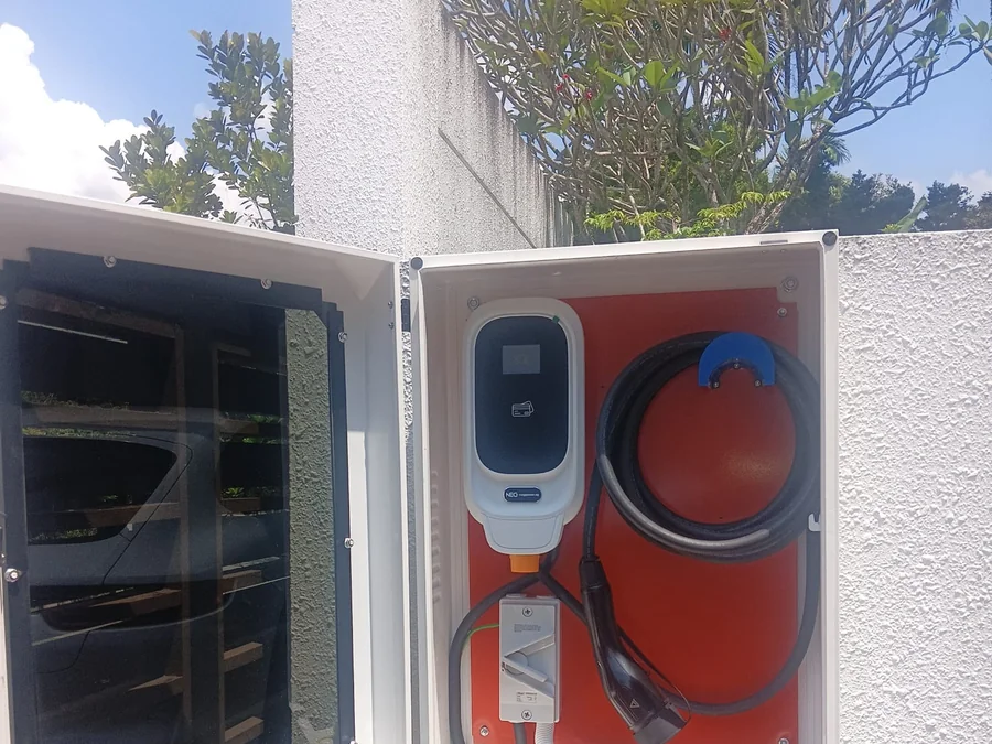 A white NEG wall charger inside an open cabinet with a red backing board, its cable coiled and the door swung wide.