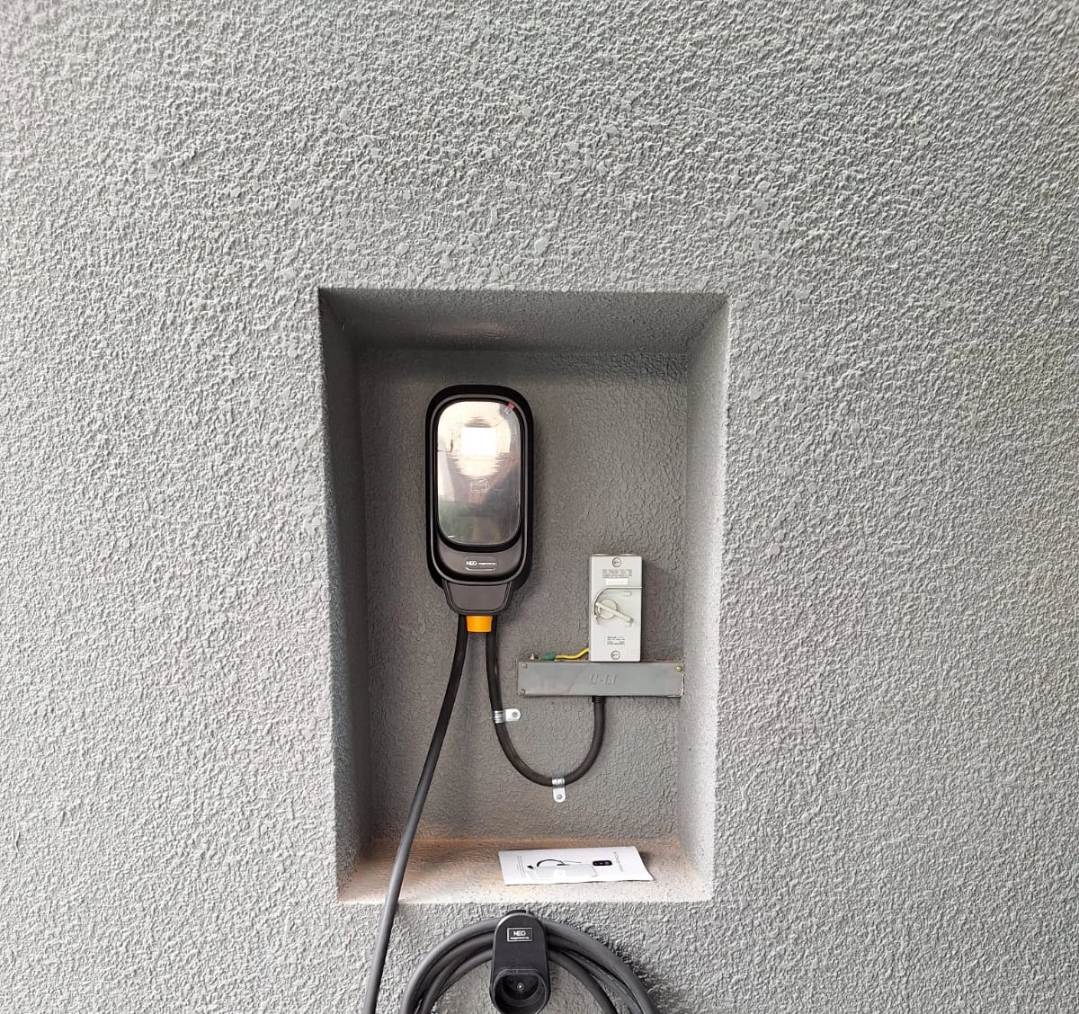 A black NEG wall charger recessed into a rendered wall niche, a small isolator beside it and the tethered connector resting on a holster below.