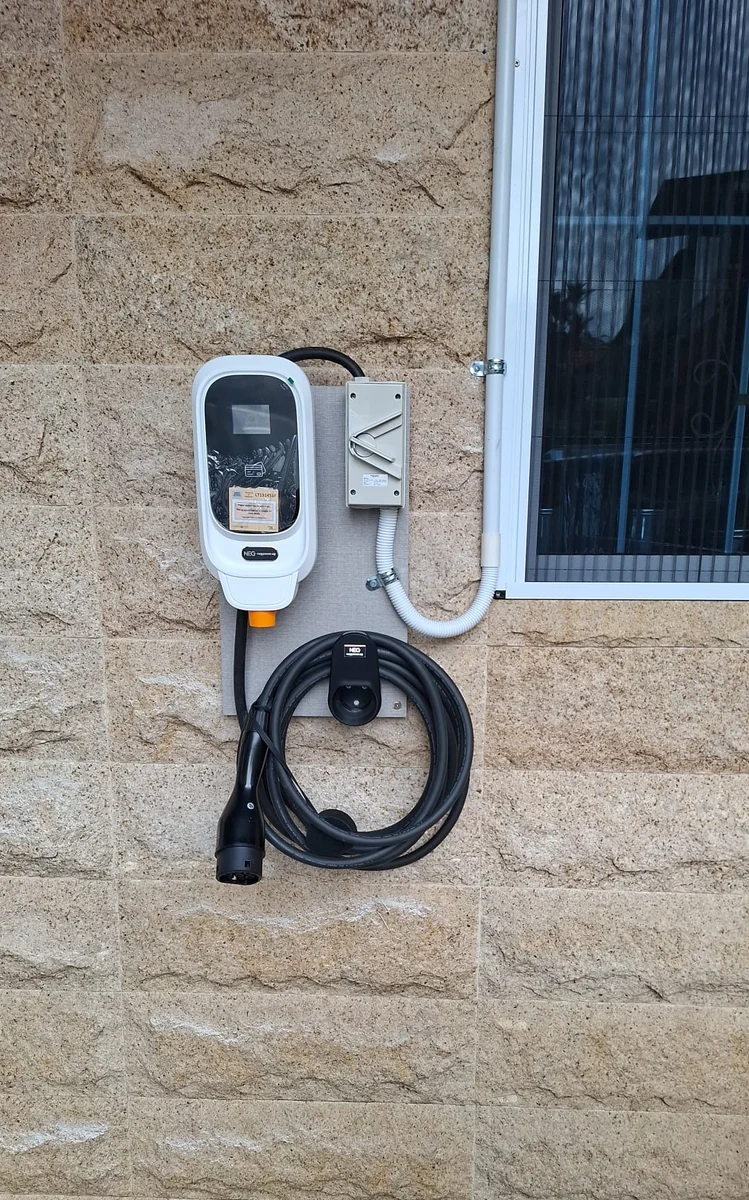 A white NEG charger on a sandstone-clad wall beside a window, its supply in white conduit and the connector coiled beneath.
