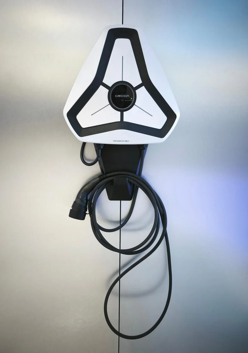 A white triangular Omoda charger on a pale wall lit with cool blue light, the cable dropping to a holster and coiling below.