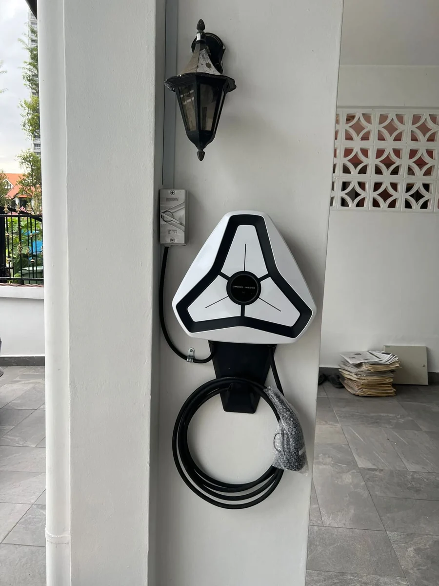 A white triangular Omoda charger on a rendered pillar with a carriage lamp above, the cable coiled on its holster and a tiled forecourt beyond.