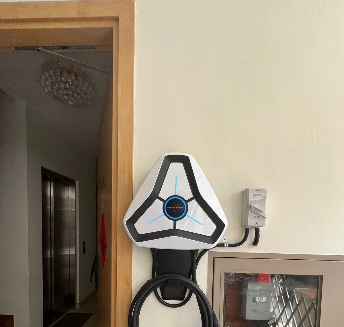 A white triangular Omoda charger on a wall in a private lift lobby, a timber door frame to the left and an isolator to the right.