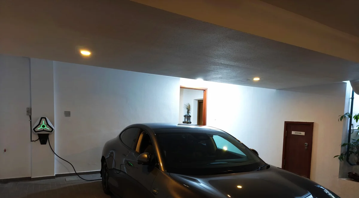 A white triangular Omoda charger glowing on the wall of a covered home garage with a dark car parked alongside and its cable running to the vehicle.
