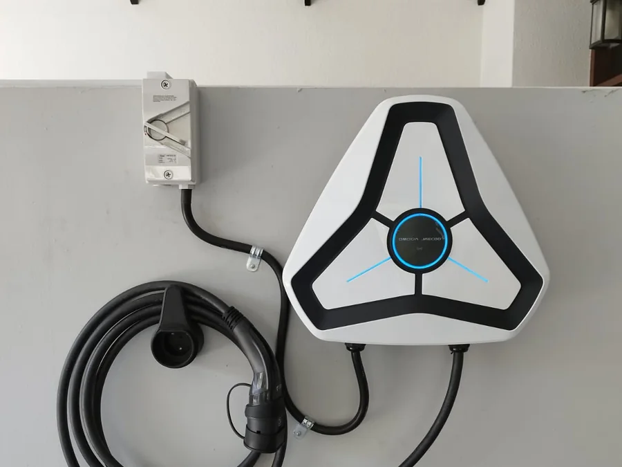 A white three-spoke charger mounted under a porch beside a lantern and decorative screening, its cable coiled beneath.