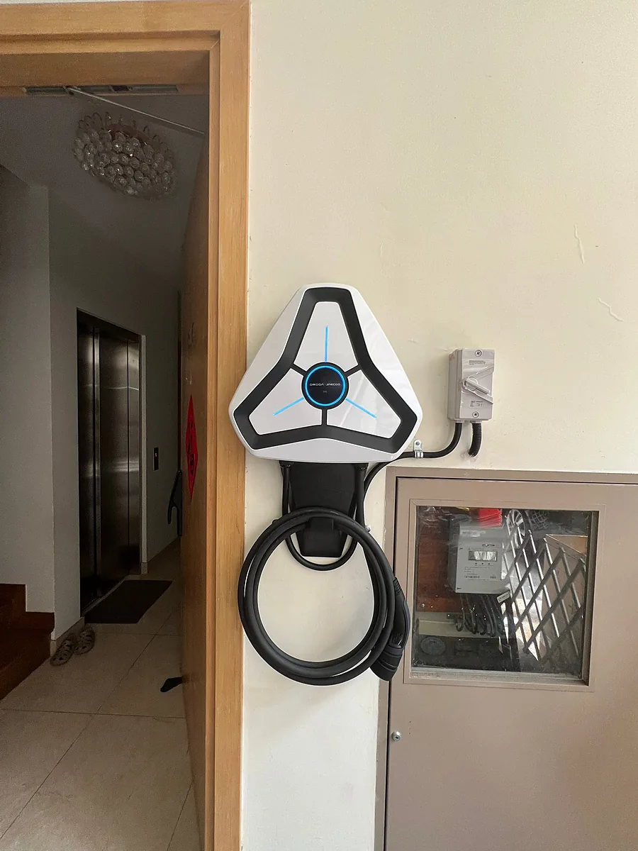 A white three-spoke charger on an interior wall beside an open doorway, its cable coiled on the holster and a glazed panel to the right.