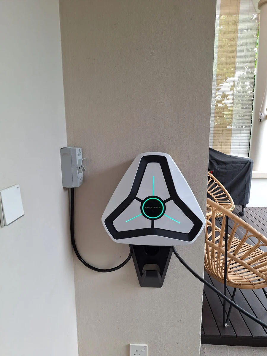 A white triangular Omoda charger on a patio wall beside an isolator, rattan furniture and a timber deck visible through the opening to the right.