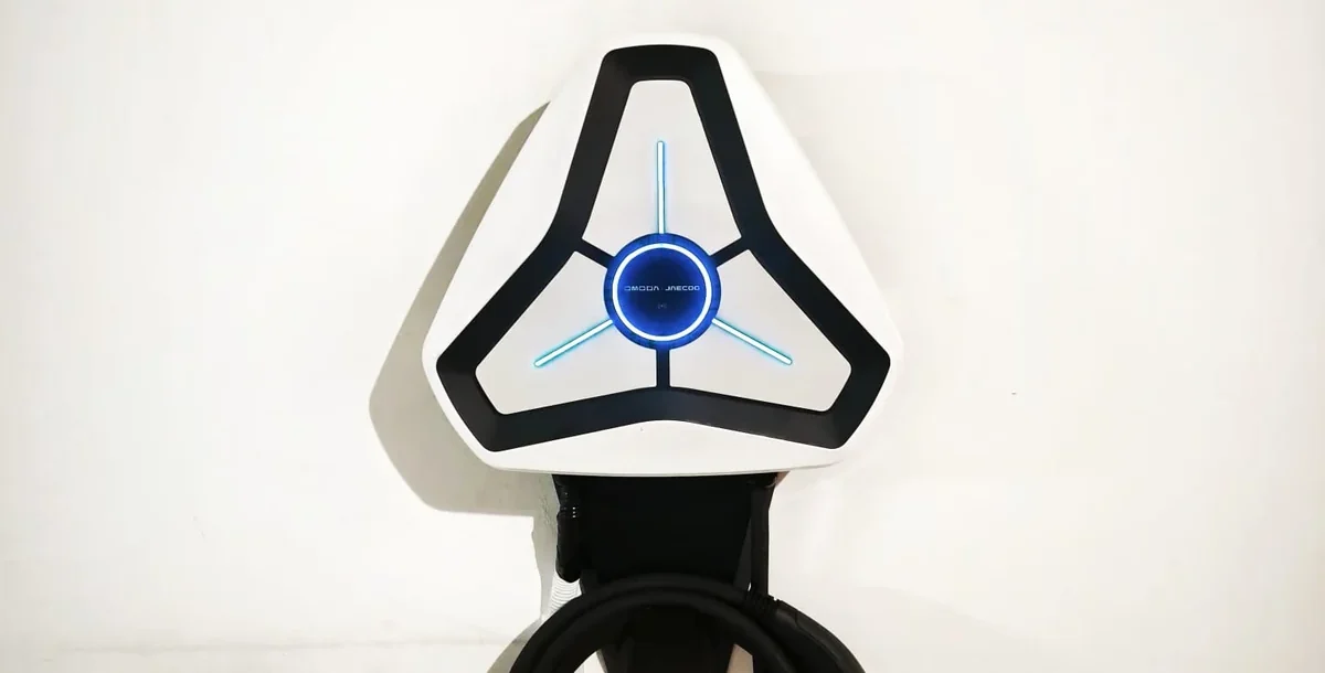 A white triangular Omoda charger with its blue illuminated ring, mounted on a plain white wall with the connector stowed beneath.