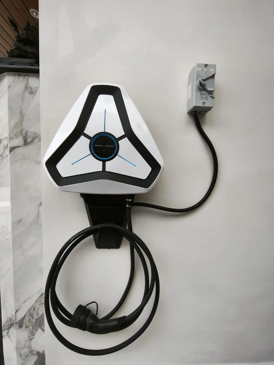 A white triangular Omoda charger on a marble-clad wall, a grey isolator to its right and the tethered cable coiled in a wide loop below.