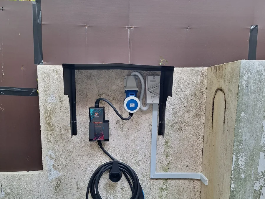 A portable charging unit mounted on a bracket on a rendered outdoor wall, the blue industrial plug and cable stowed beneath.