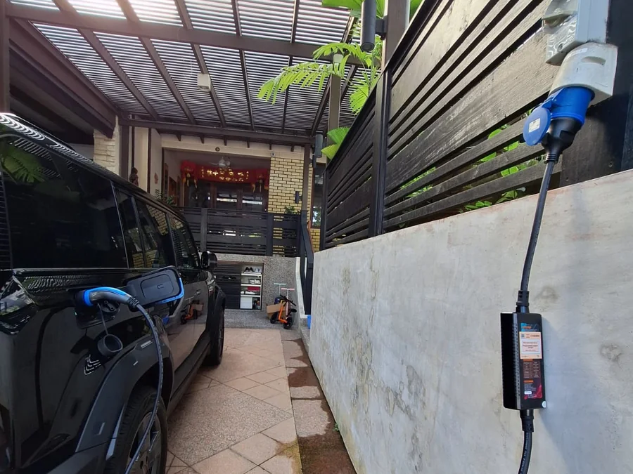 A portable charging unit hanging on a driveway wall with the cable running to a vehicle parked alongside, under a covered carport.