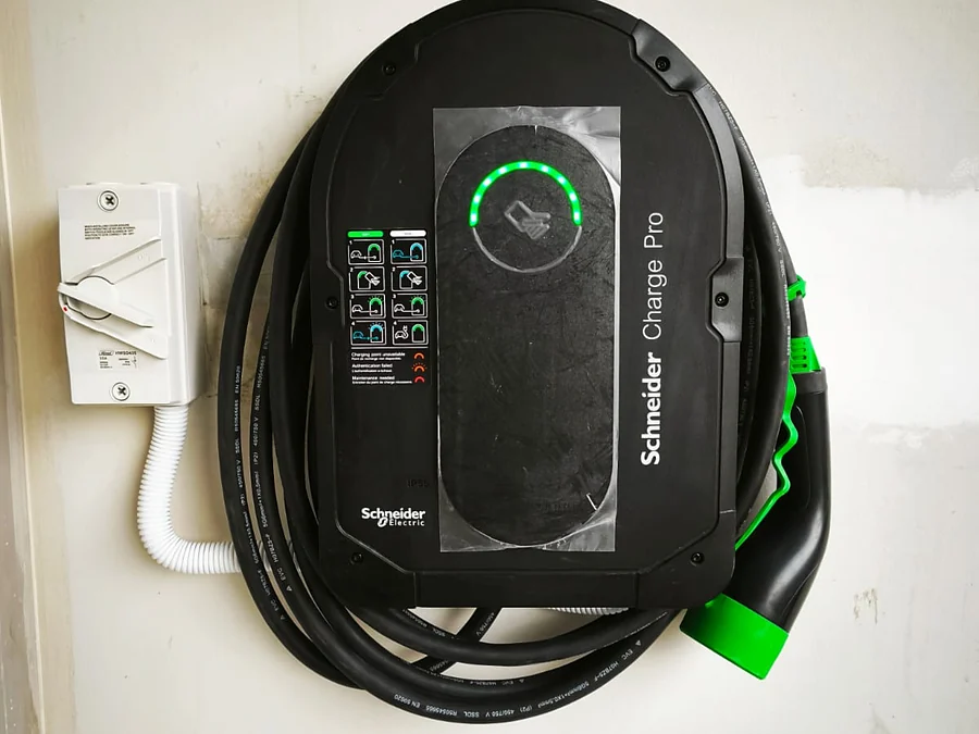 A black Schneider Charge Pro on an interior wall with its status ring lit green and the protective film still on the fascia, an isolator switch to the left and the tethered cable coiled with its green connector.