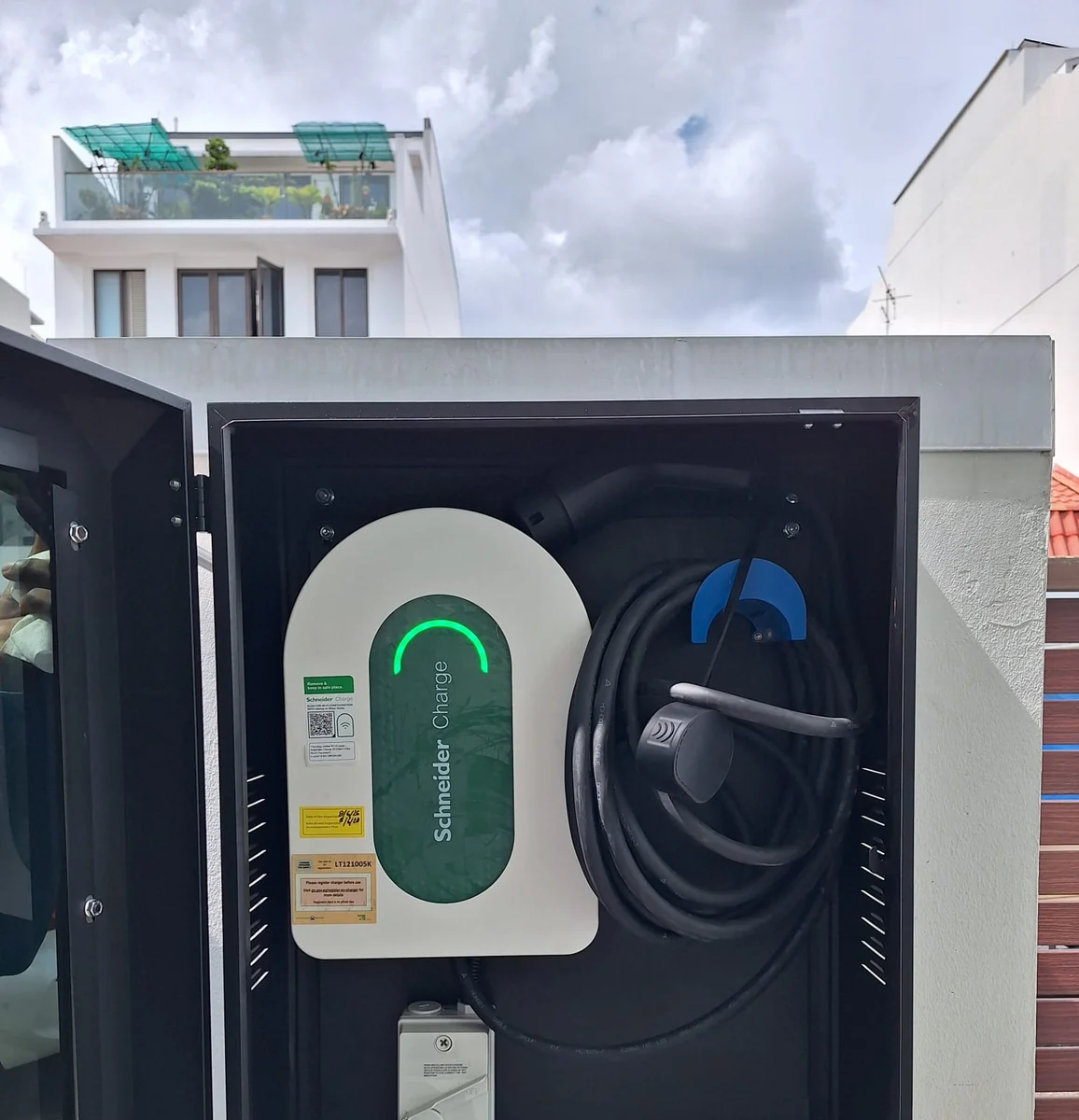 A white Schneider Charge unit inside an open lockable steel cabinet mounted on a brick wall, its status ring lit green and the blue-tipped connector stowed alongside.