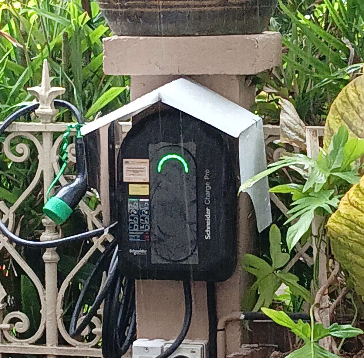 A black Schneider Charge Pro with its status ring lit green, mounted on a masonry pillar under a folded metal weather hood, wrought-iron railings and dense planting either side.
