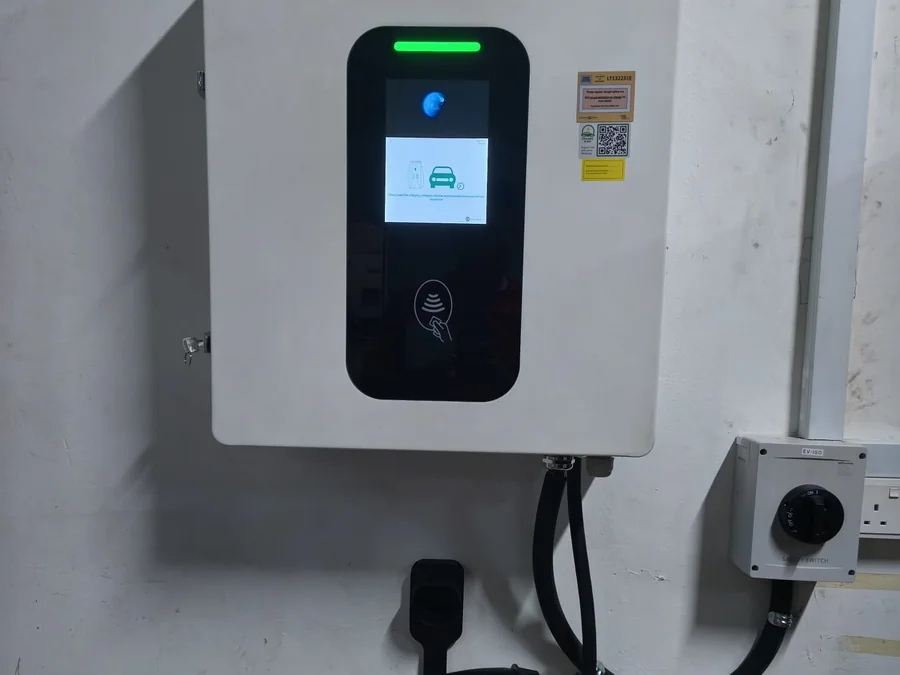 A Sinexcel wall-mounted charging unit with a colour display and card reader, mounted on an exterior wall with its cable coiled below.