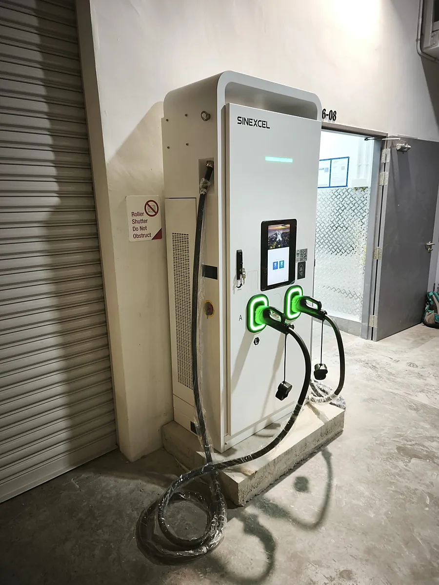 A white floor-standing DC EV charger on a concrete plinth indoors, its two connectors holstered in green-lit surrounds, with a plastic-wrapped supply cable running from an opening in the wall and coiled on the floor beside it.