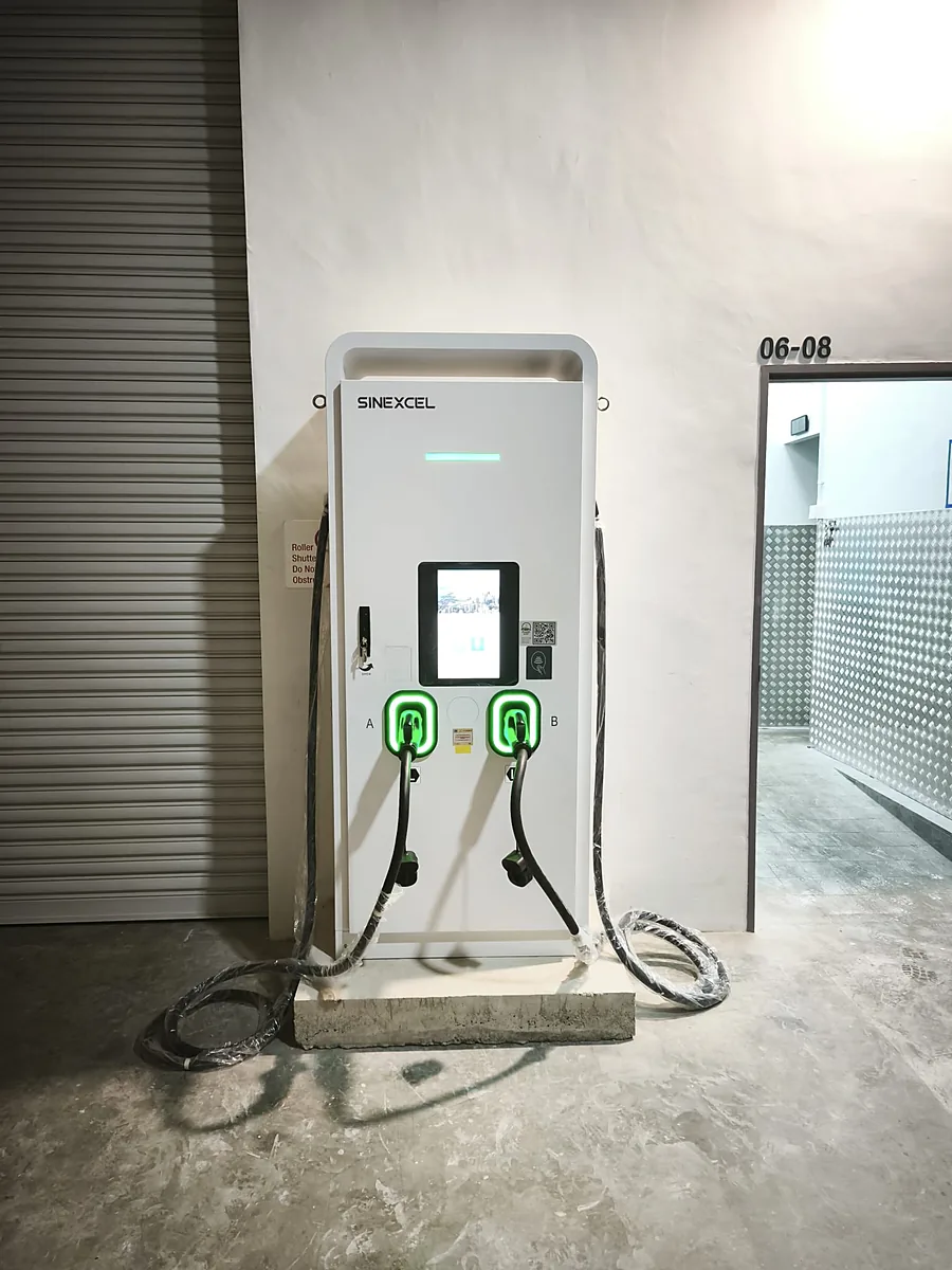A white floor-standing DC EV charger seen straight on, its two holstered connectors ringed in green light and their wrapped cables looping to the floor.