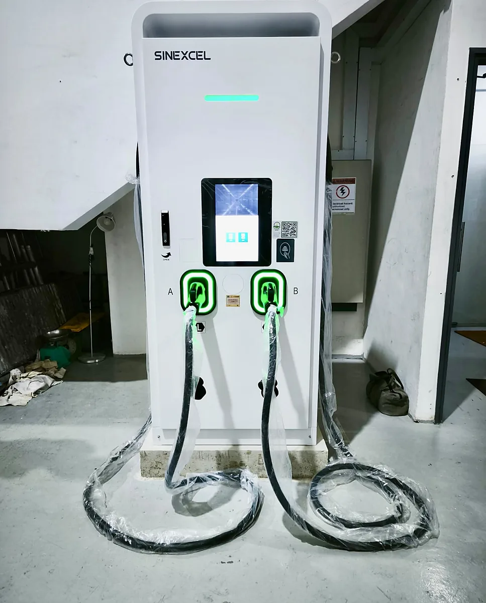 A white floor-standing DC EV charger on a concrete plinth indoors, its two holstered connectors ringed in green light and their wrapped cables looping across the floor.