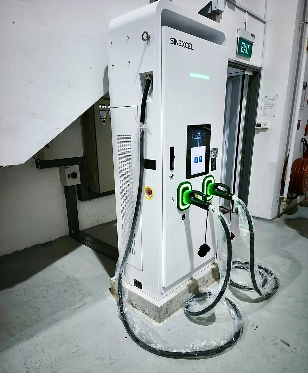 A white floor-standing DC EV charger on a concrete plinth indoors, both connectors seated in their green-lit holsters with their plastic-wrapped cables looping down to the floor.