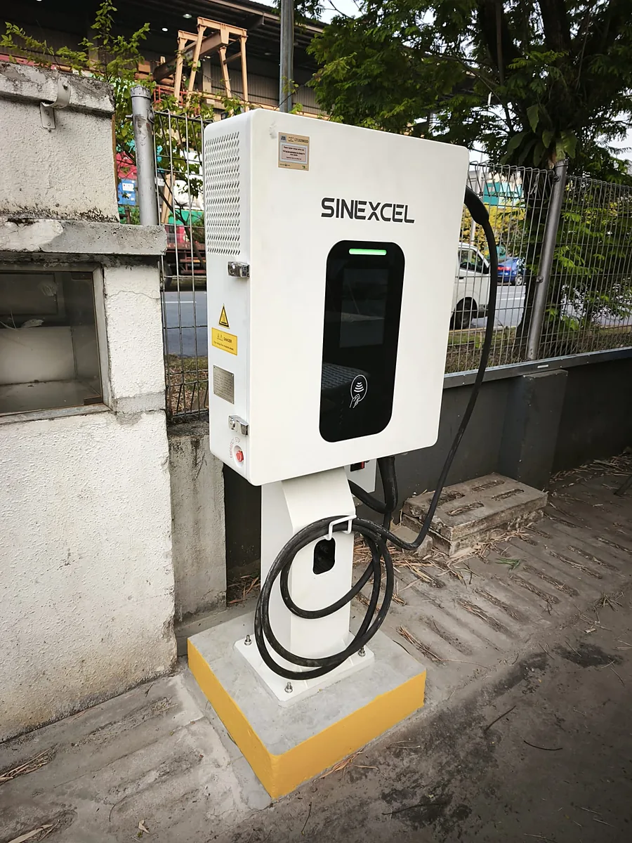 A white pedestal-mounted EV charger with a dark screen panel and a green status bar, its black cable coiled around the pedestal and hung on a bracket, bolted to a yellow-edged concrete plinth beside a mesh fence.