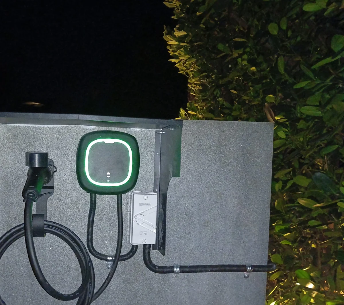 A dark Wallbox unit with its ring lit white on a rendered garden wall at night, an isolator and flexible conduit to its right and the tethered cable coiled on a holster to the left.