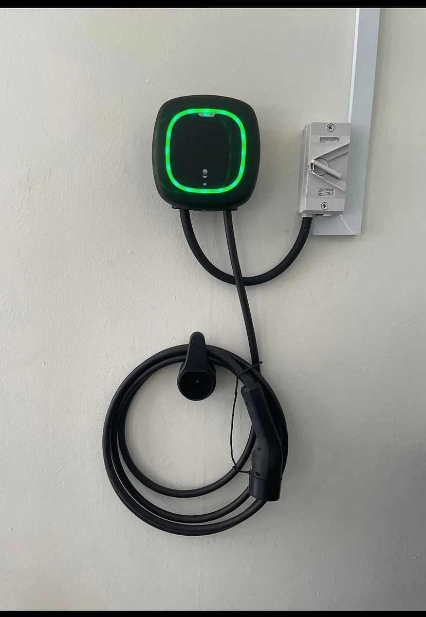 A dark Wallbox unit on a white interior wall with its status ring lit bright green, a white isolator switch beside it and the cable coiled neatly on its holster below.