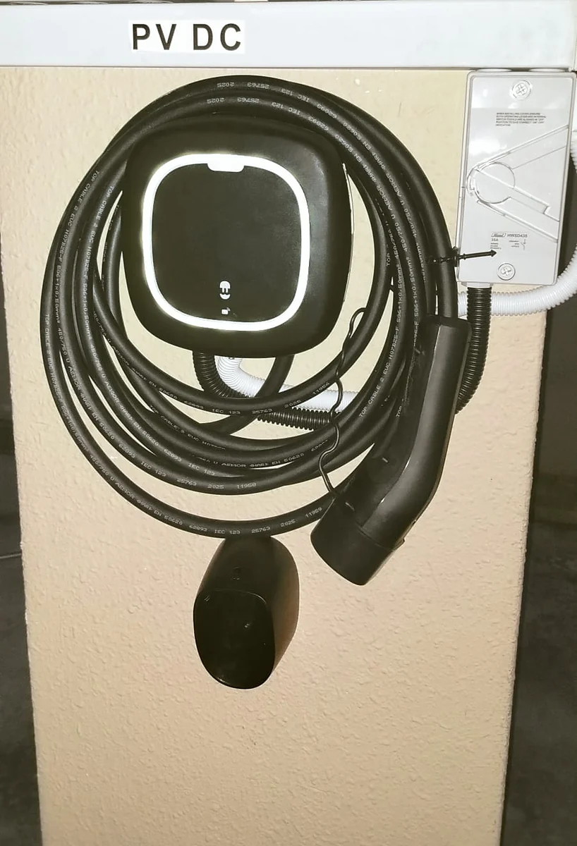 A dark Wallbox unit mounted on a pillar with the tethered cable coiled in a wide loop around it, an isolator to the right and a distribution panel above.